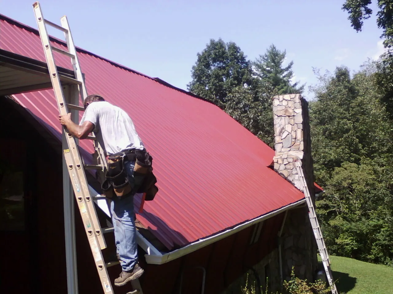Roofer on a ladder beside a red metal roof and stone chimney — Soffit & Fascia Repair by Roofing Brothers in Clovis